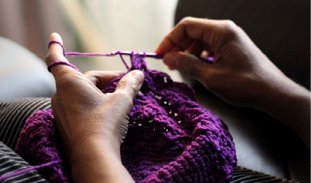 a Black woman's hands crochet a purple yarn craft