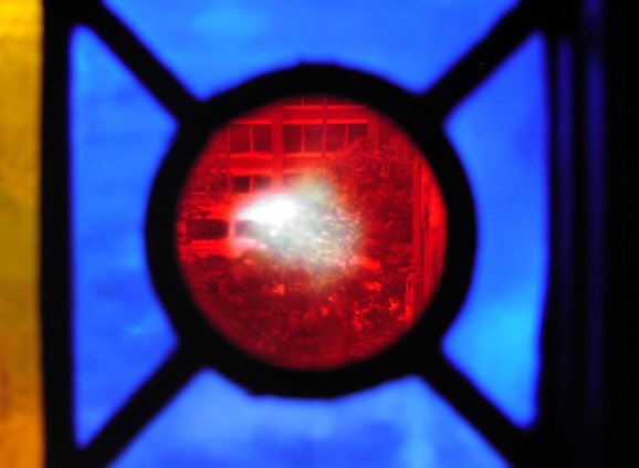 Stained glass in cobalt blue, golden orange, and red. There is a red circle surrounded by four blue almost triangles, with an orange bar on the left-hand side.