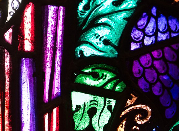 A stained glass detail showing purple grapes and vivid green leaves. There is a yellow vine curl as well as purple and red folds of stained glass.
