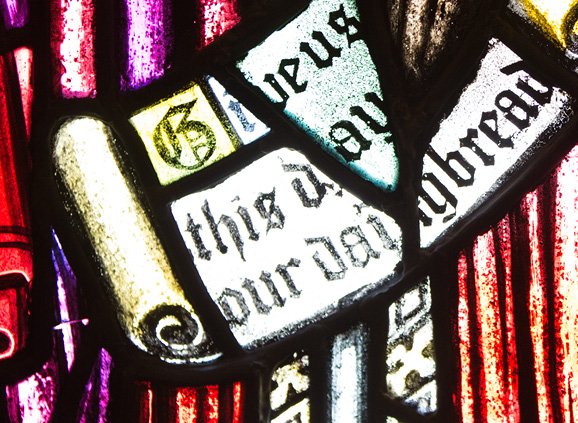 purple, red, and white stained glass depicting a scroll. The scroll reads in Gutenberg-style text: 'Give us this day our daily bread'