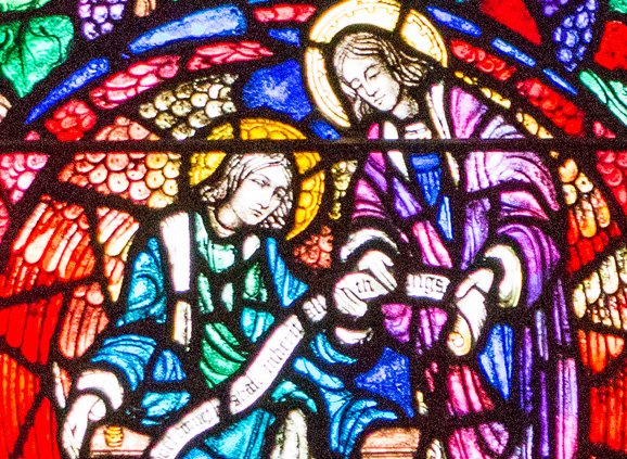 Two angels hold a scroll between them. The angels are made up of brilliant pieces of stained glass in green, blue, purple, with yellow and red in their wings and purple grapes and green leaves surrounding them.