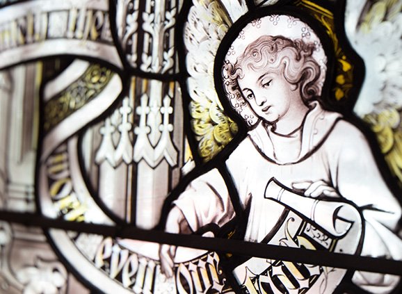 A mostly black & white image of a stained flass window of a winged angel with caucasian features holding a scroll. The legible section of the scroll reads 'even our faith' in a fancy calligraphy hand.