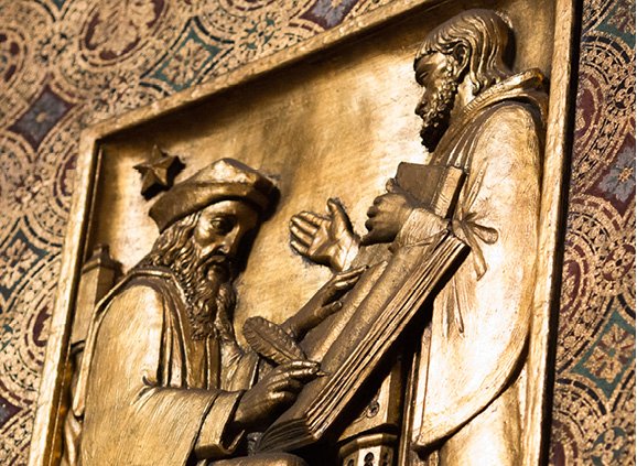 In gold relief, a seated man with a long beard is writing with a feather pen into a book. Another man, with a short beard and holding a book, is talking to him. A circular pattern in primarily gold, red, and green, surrounds the relief image. 