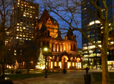 Trinity Church Boston glows on Copley Square as it is wrapped in Christmas lights and a Christmas tree is in the foreground. The sky is a French blue as it is dusk.