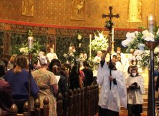 Photo of an Easter procession with people, a silhouetted cross, and flowers down the center aisle of Trinity Church Boston (Episcopal) in Copley Square.