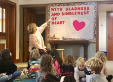 a teacher in a classroom points to a bulletin board with the words 'gladness and singleness of heart' in pink letters, with a big pink heart. The backs of children who are sitting on the floor can be seen watching the teacher.