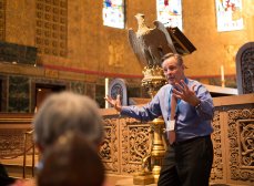 A tour guide enthusiastically talking to several people in the pews of Trinity Church Boston, with the golden chancel and the eagle lectern behind the tour guide