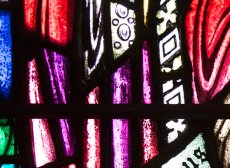 Colorful stained glass that looks like folds of cloth in cobalt blue, violet, and red, with patterned black and white blocks breaking up the space about two-thirds of the way into the image. 